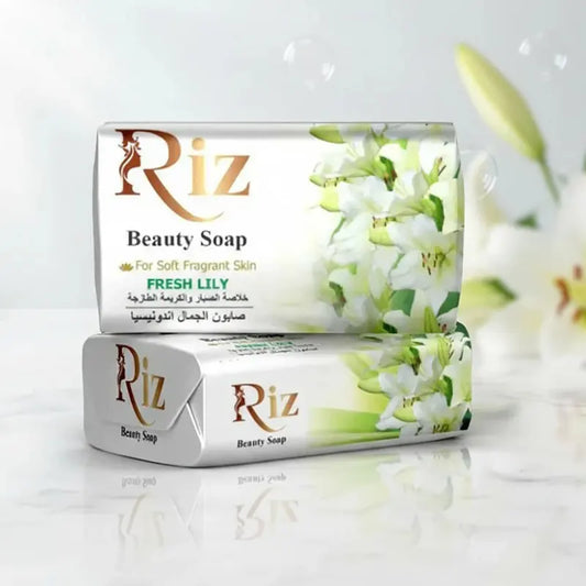 Two bars of Riz Beauty Soap with "Fresh Lily" scent stacked on a white marble surface. The packaging is white with elegant gold lettering and illustrations of blooming white lilies. In the background, soft-focus lilies and floating soap bubbles create a clean, refreshing atmosphere.