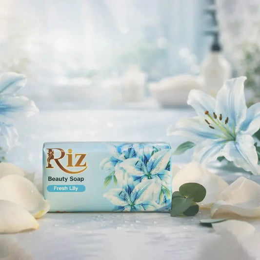 A bar of Riz Fresh Lily Beauty Soap in light blue packaging, positioned centrally on a textured surface. The scene is surrounded by soft white lily petals and elegant light blue lily flowers against a bright, airy, and blurred background.